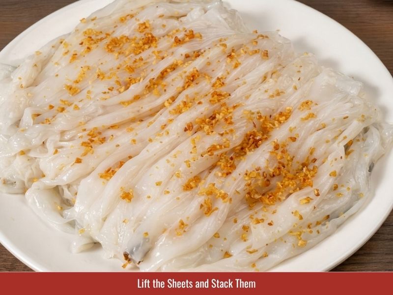 Removing cooked banh uot rice sheets and stacking them with shallot oil to prevent sticking