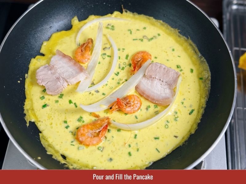 Pouring thin banh xeo batter into a hot skillet and adding pork, shrimp, and bean sprouts