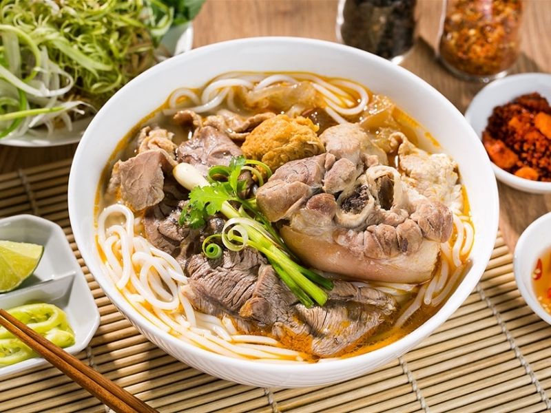 Bowl of bun bo Hue with sliced beef, pork hock, thick rice noodles, spicy lemongrass broth, and fresh herbs